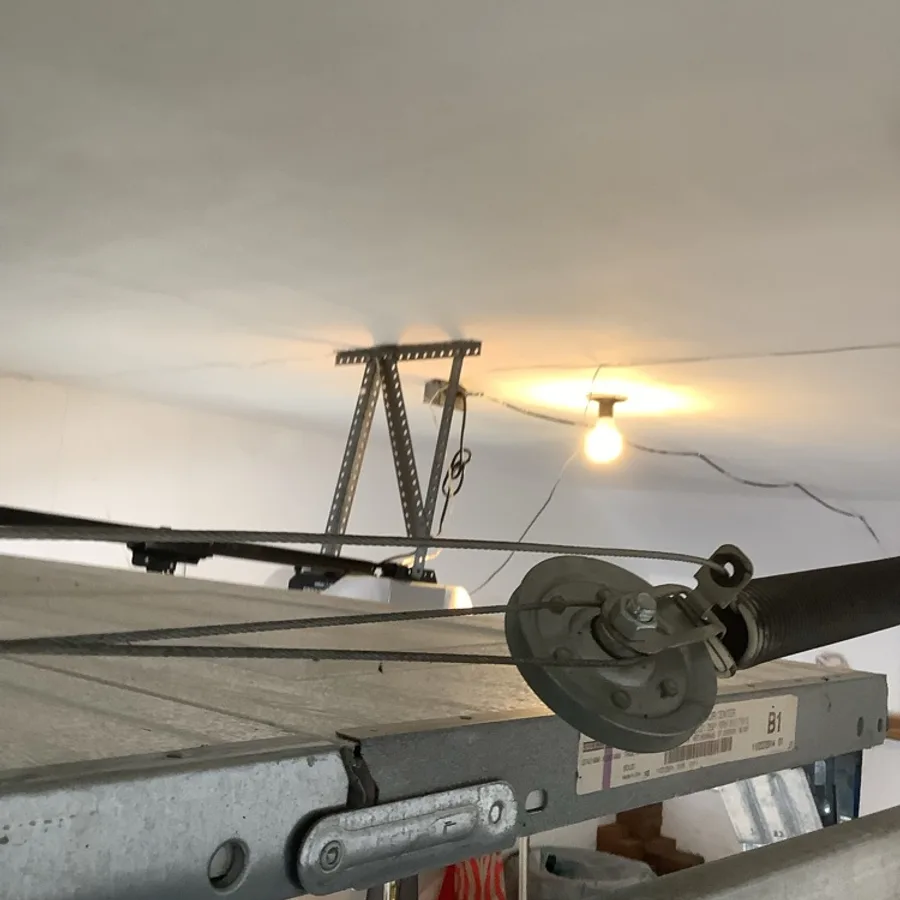 A cable drum and two taut lift cables at the top corner of a garage door, with the opener rail bracket and a bare ceiling bulb behind them.