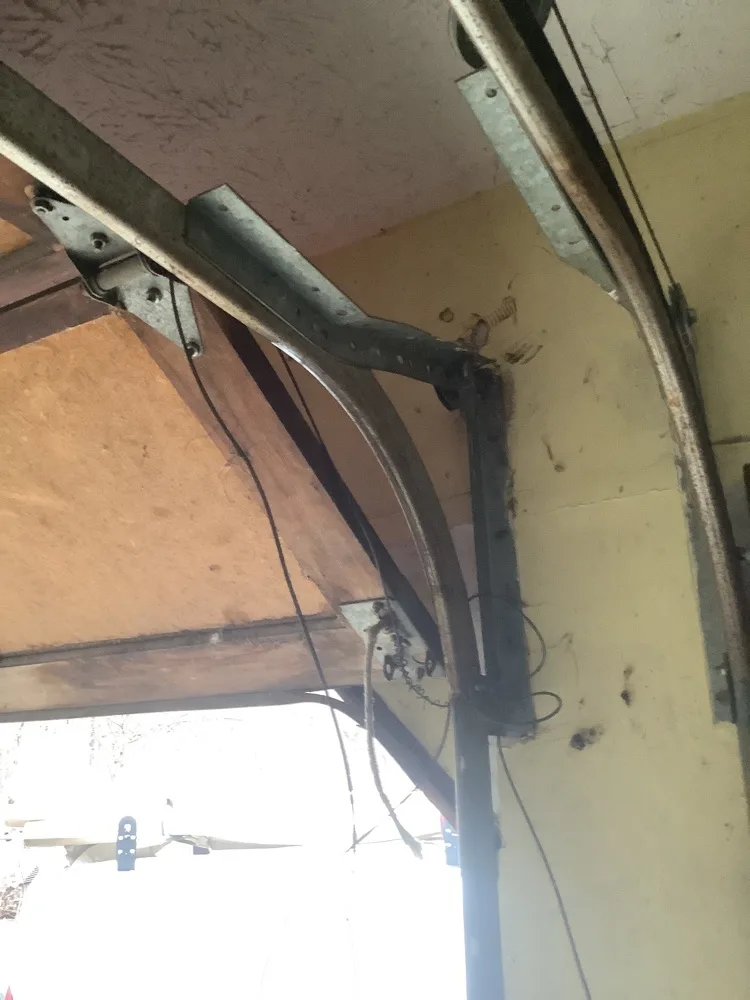 The radius bend where a vertical garage door track curves into the horizontal, held by a punched steel bracket, with the spring and cable behind it against a yellowed wall.