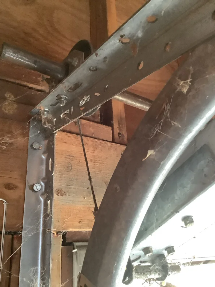 The curved section of an old galvanised garage door track bolted into rough timber framing, a small pulley behind it and cobwebs strung between the members.