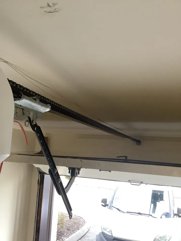 A chain-drive opener rail and its angled door arm seen from inside a garage, the open door above and a white work van parked on the driveway beyond.