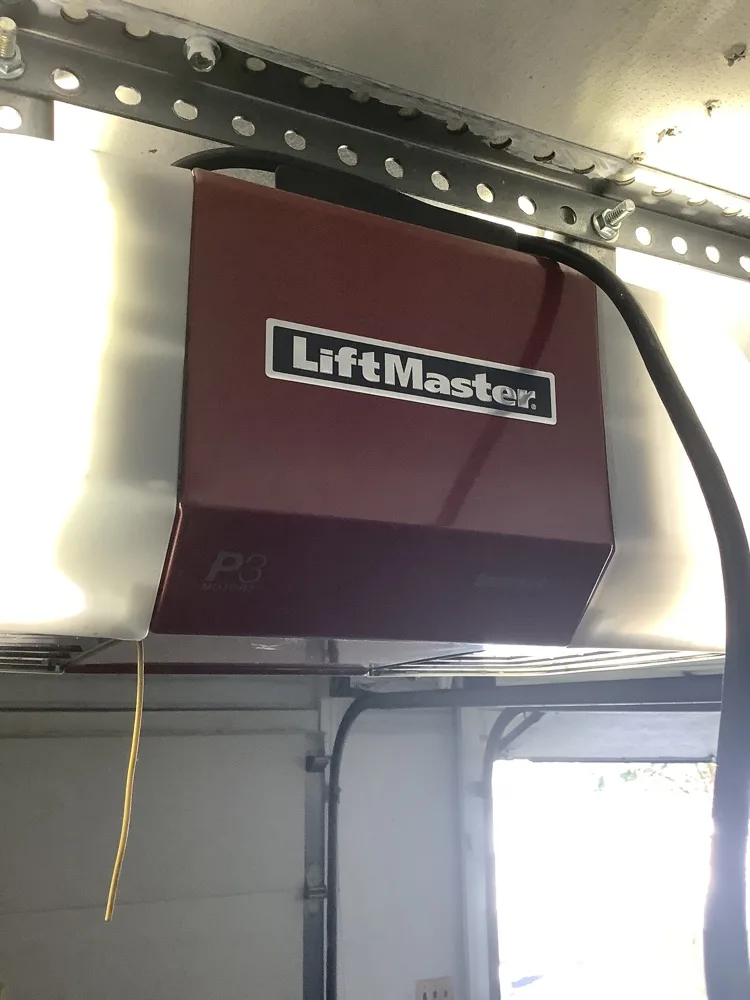 A maroon LiftMaster P3 opener head bolted to punched angle iron above a garage door, its light lens glowing at the side and a pull cord hanging from the housing.
