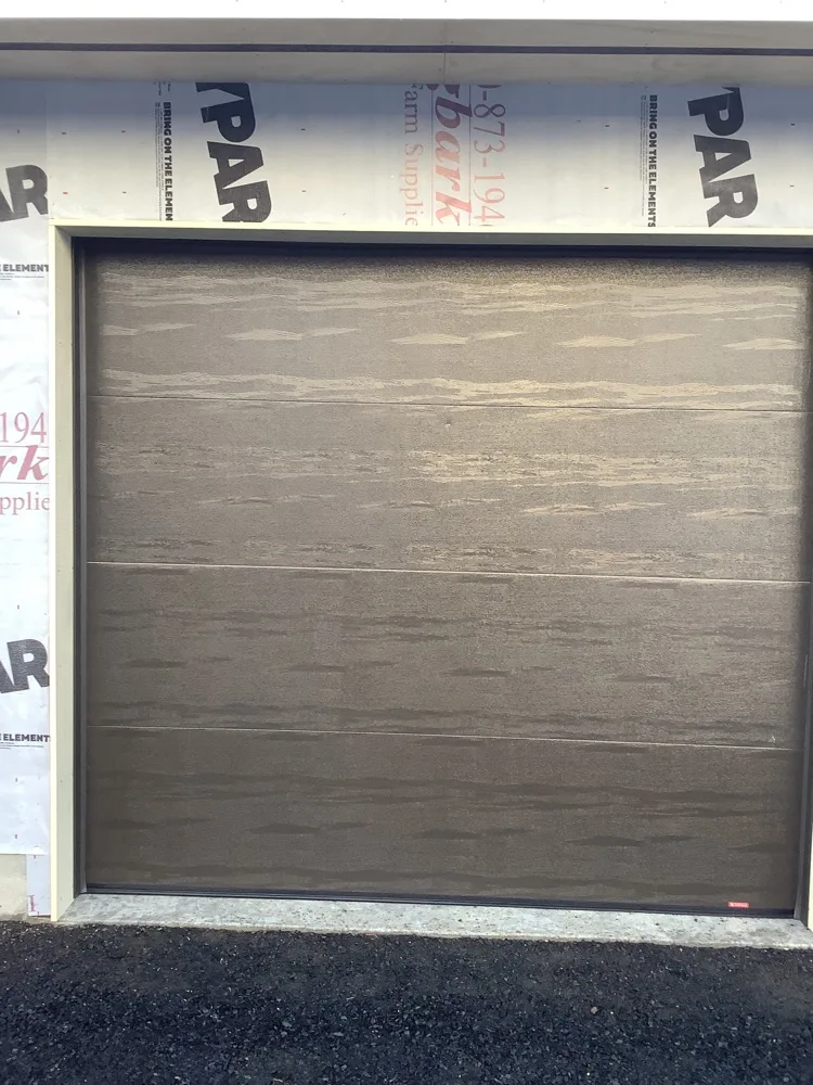A newly fitted garage door in a bronze woodgrain finish sitting square in its opening, the surrounding wall still wrapped in white housewrap and the concrete apron freshly poured.