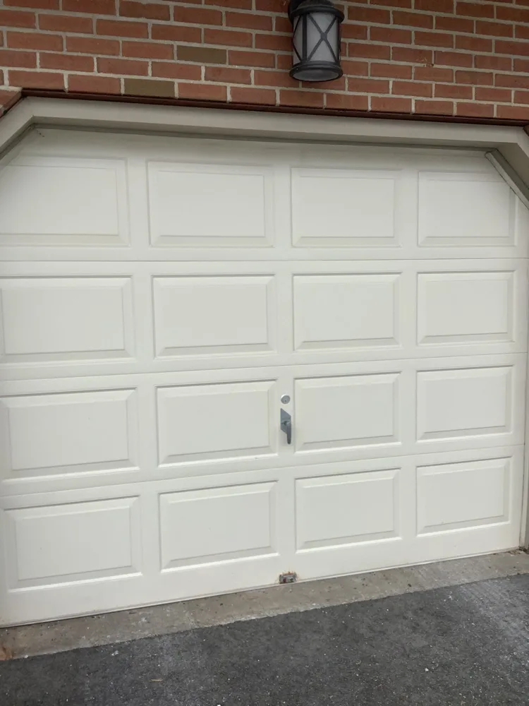 A cream raised-panel garage door in good order beneath a red brick wall, with a black coach lamp mounted above the opening and a clean concrete apron below.