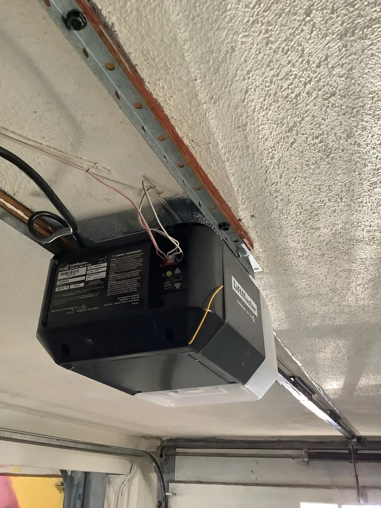 A black LiftMaster myQ opener head seen from below against a heavily textured ceiling, its wiring looped over the rail and the top of the door visible at the bottom of the frame.