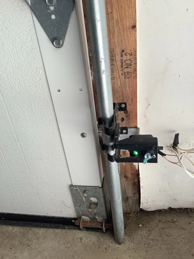 The bottom of a garage door track where it meets the concrete, showing the jamb bracket, the closed door edge against the floor and a safety sensor showing a green light on the timber jamb.