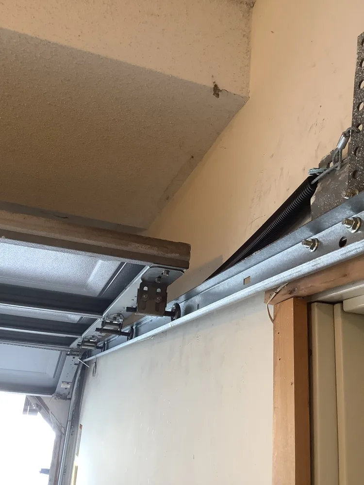 The horizontal track of a garage door running back from the radius under a sand-coloured ceiling, carried on bolted hangers with an extension spring stretched alongside it.