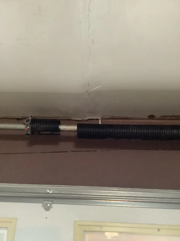 A black torsion spring wound onto its shaft across a garage header, the winding cone at the near end and a long crack running through the plaster ceiling above it.