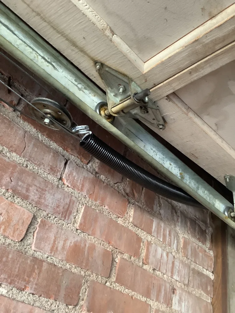 An extension spring hooked to a steel pulley at the end of a garage door track, mounted against an old red brick wall with the lift cable running back over the sheave.