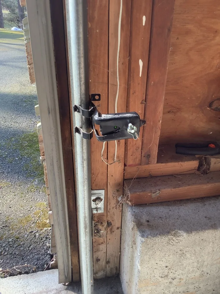 A safety photo eye clamped to the vertical track on a timber jamb, its indicator lit green, with the gravel driveway visible through the open doorway beside it.